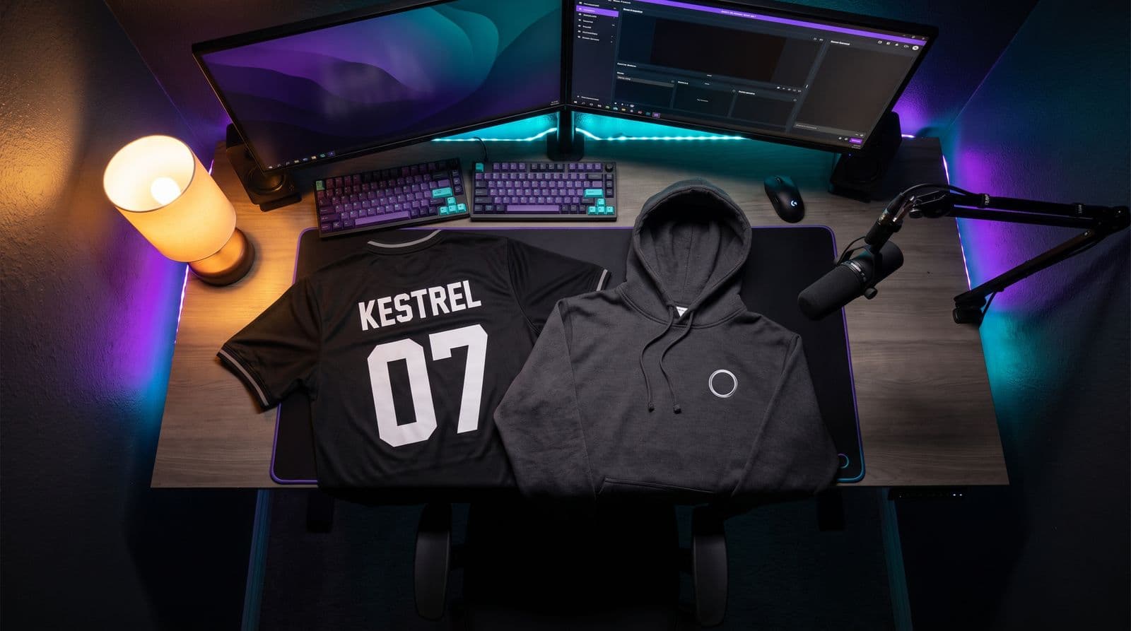 A black esports jersey turned to show KESTREL 07 on the back and a charcoal hoodie with a small ring emblem, laid across a streaming desk lit by purple and teal LEDs.